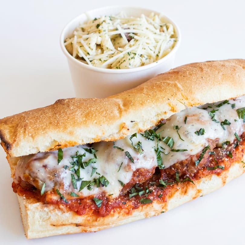 Meatball sub sandwich with melted cheese and parsley, next to a cup of coleslaw.
