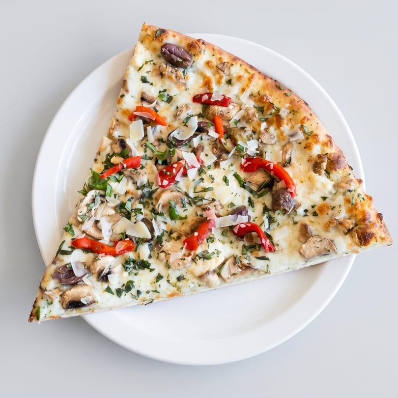 A large slice of white pizza with mushrooms, red peppers, olives, and herbs on a white plate.
