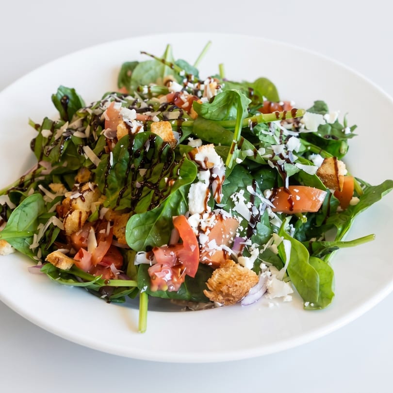 Spinach salad with tomatoes, feta cheese, croutons, and balsamic glaze on a white plate.